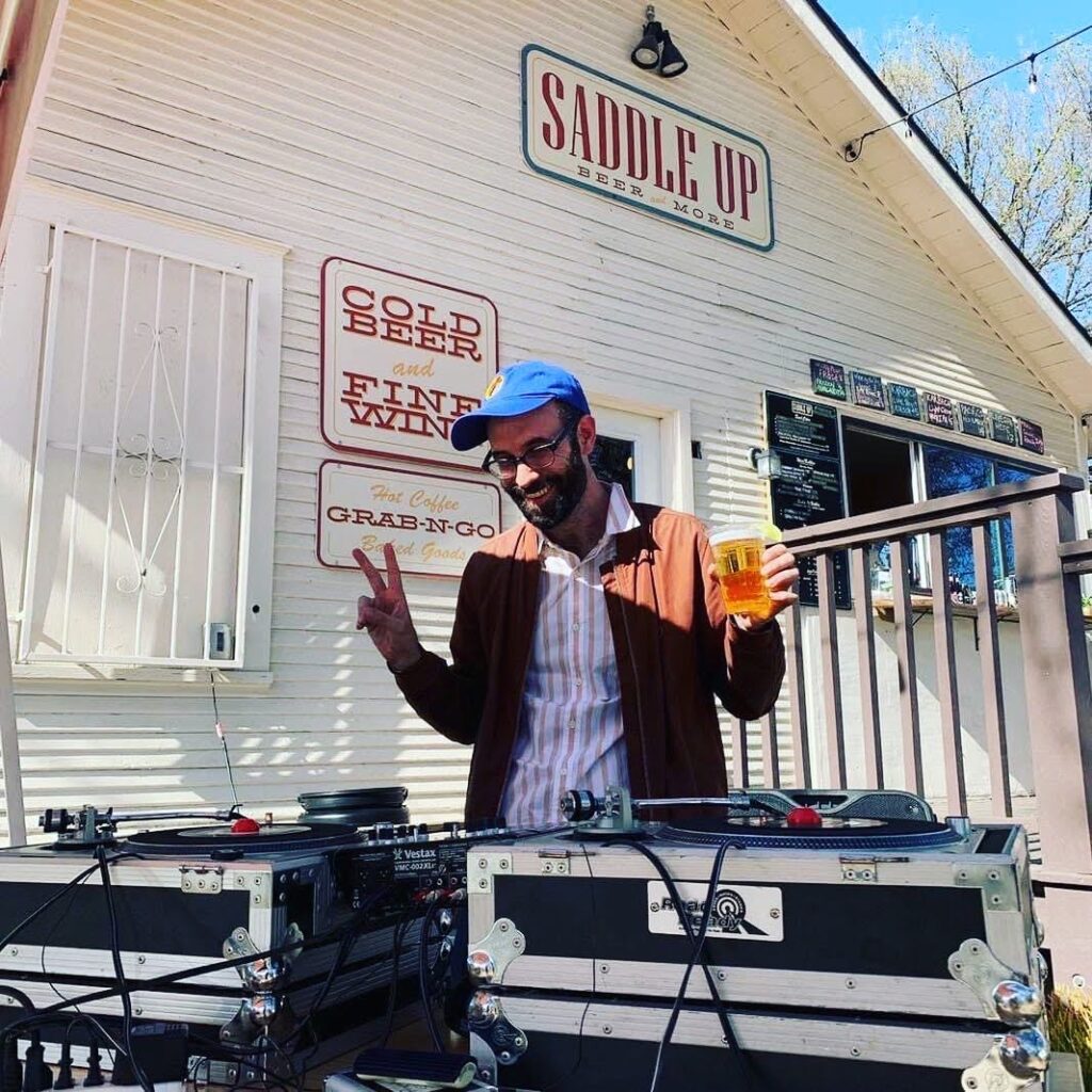 dj making hand sign gesture holding beer, in front of two turntables, outdoors